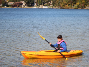 A woman in a blue jacket paddling an orange kayak on a lake.